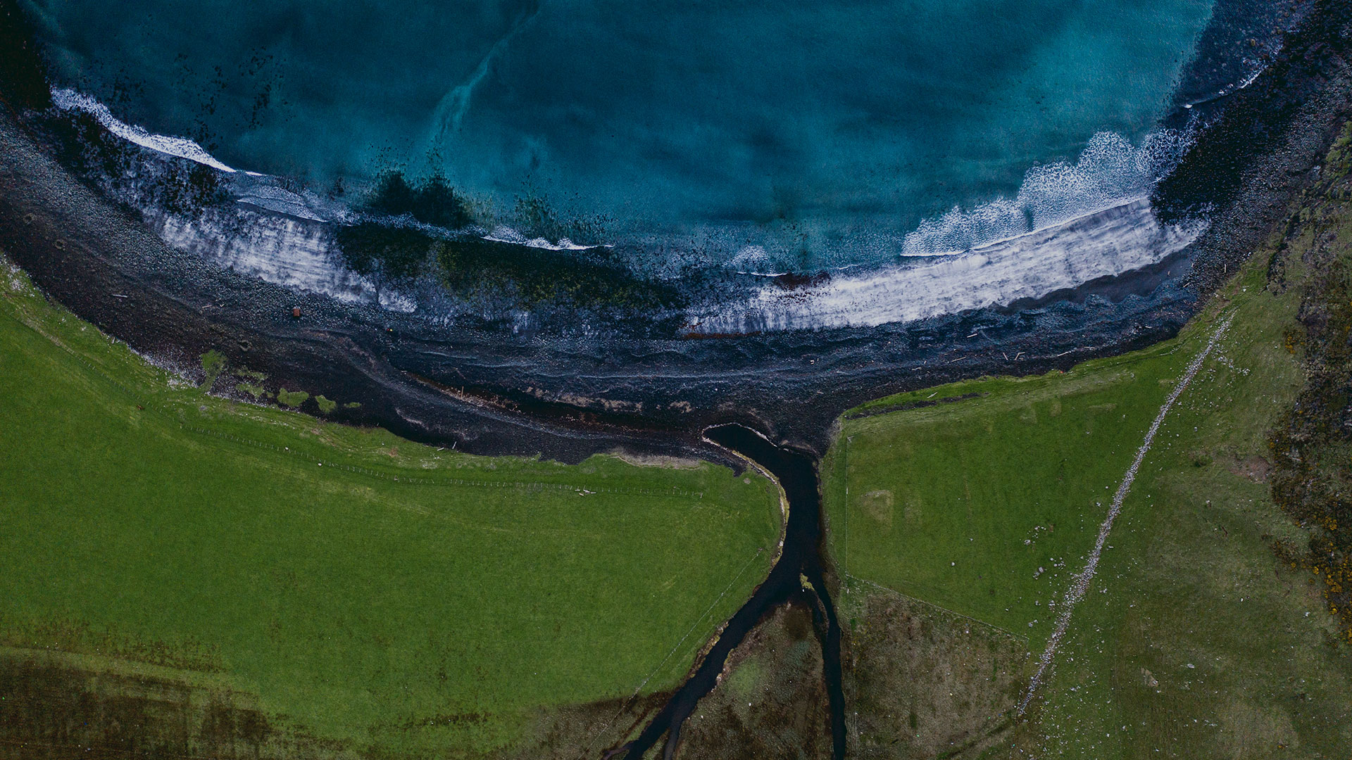 Overhead perspective of a lush green field adjacent to the calm ocean in Scotland, highlighting the beauty of nature's contrast.